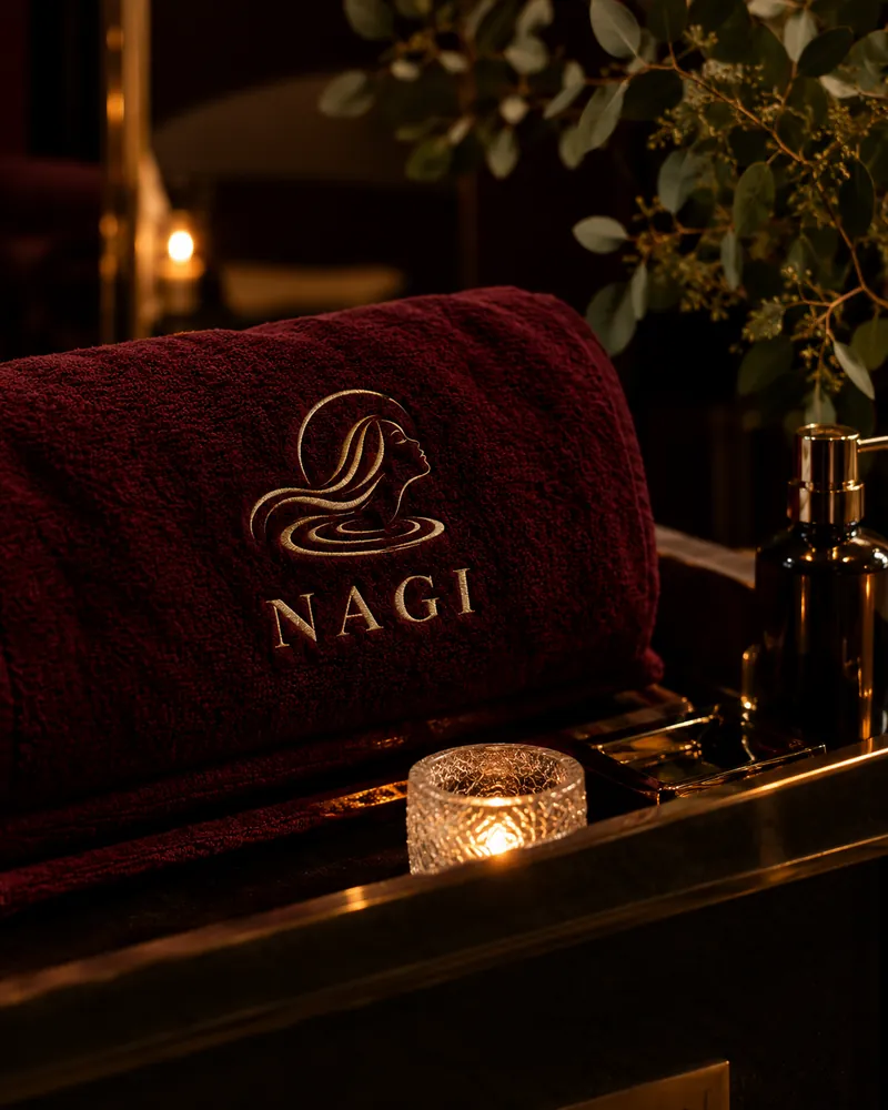 A folded burgundy NAGI towel beside a glowing candle at the basin